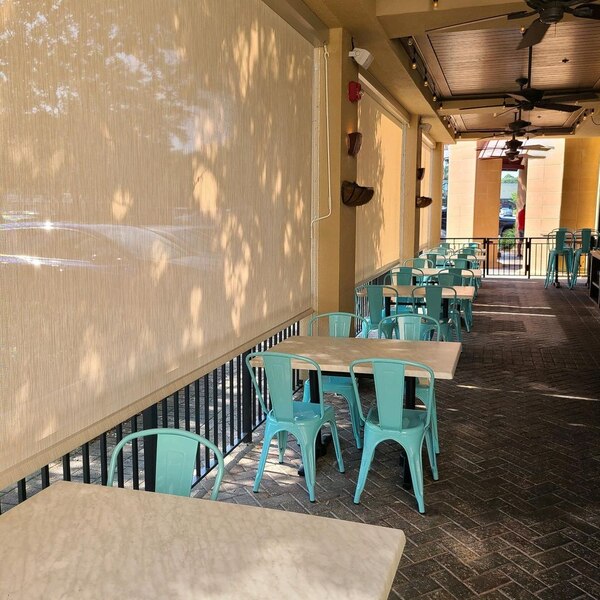 Roll down solar shades, auqua colored metal chairs and beige tables line a metal and stucco outdoor patio area of a restaurant.